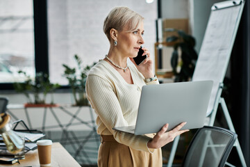 A middle-aged businesswoman with short hair talks on a cell phone while holding a laptop in a...