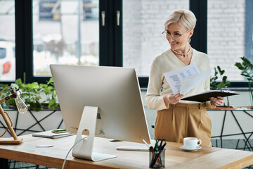 A middle aged businesswoman stands in front of a computer, holding a piece of paper while working in a modern office setting.