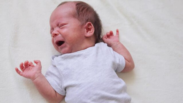 Infantile colic. Baby care. Close up of crying newborn laying on bed wearing bodysuit sad hangry baby screaming loud while being alone in bedroom