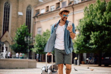 Happy man with travel bag text messaging on cell phone while walking in  city.