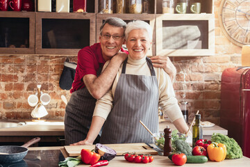 A senior couple smiles while preparing a meal in their kitchen. The woman is leaning forward, her hand resting on the countertop. There are fresh vegetables and a bowl of salad on the counter