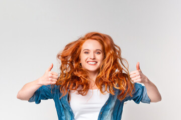 A Caucasian woman with vibrant red hair, wearing a casual outfit, is smiling and making a thumbs up...