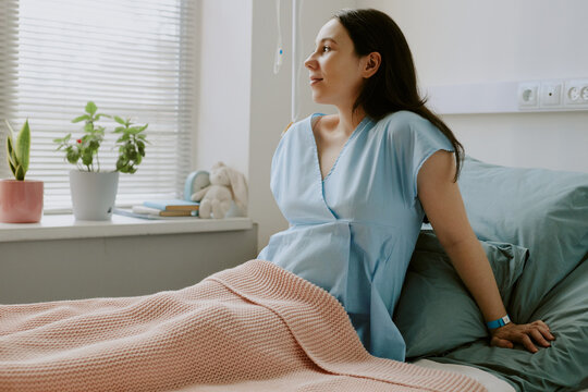Young pregnant woman sitting on bed and looking out of window while she staying at maternity hospital