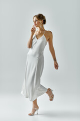 A young, beautiful blonde woman gracefully walks in a flowing white dress against a soft grey...