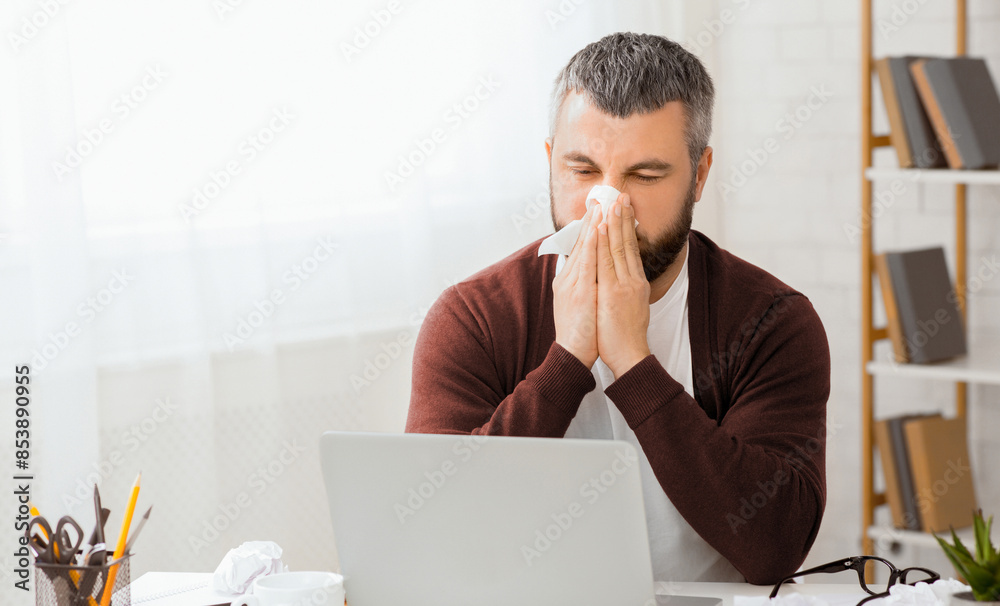 Aufkleber Sick man sitting in front of a laptop is blowing his nose into a tissue. He appears to be dealing with a cold or allergies while using the computer.