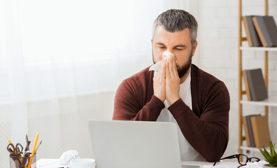 Sick man sitting in front of a laptop is blowing his nose into a tissue. He appears to be dealing with a cold or allergies while using the computer.