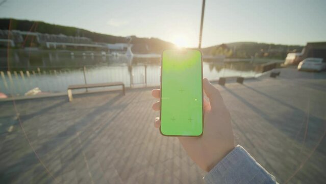 At sunset hands holding vertically phone smartphone device with green screen moving around. On background beautiful lake. Nature