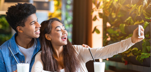 African American young guy and girl are sitting at a table in a cafe, holding a smartphone up to take a selfie together. The cafe is adorned with modern decor