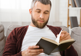 A man is seated on a couch, engrossed in reading a book. He is focused and turning the pages, with...