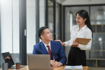 Young professionals collaborating in an office environment. A woman stands while discussing work with a seated man at a desk with a laptop.