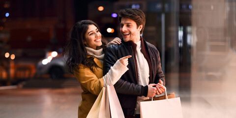 Look here! Woman showing man fashion clothes in shop window, walking in the evening near shopping centre