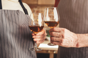 Cropped of senior couple stands in their kitchen, both wearing aprons and smiling at the camera. They are holding wine glasses and toasting each other.