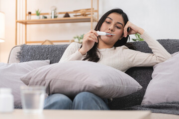 Obraz premium Young Woman Checking Temperature with Thermometer While Sitting on Couch at Home
