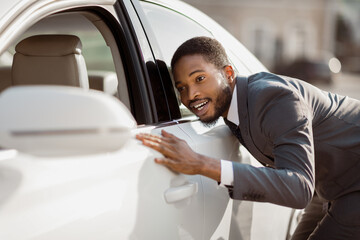 African American man in a suit looking at a new car with a look of admiration. He is standing next to the car, and his hand is resting on the door. The car is white and appears to be brand new