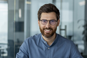 Obraz premium Close-up portrait of a young smiling man in glasses and a blue shirt sitting and working in the office and looking confidently at the camera