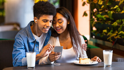 Two African American teenagers are sitting at a cozy cafe, enjoying milkshakes and a slice of cake. They are smiling and watching something on a smartphone together.