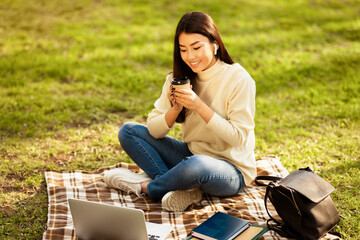 Asian young woman sits on a plaid blanket in a grassy park, sipping coffee and studying with a...