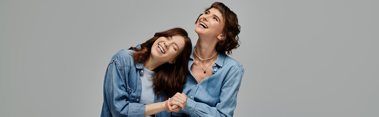 Two young women, dressed in denim, laugh together against a grey backdrop. Lgbt couple