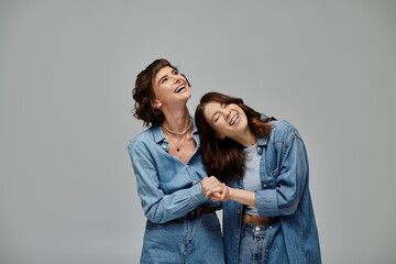 Two young women stand next to each other, arms wrapped around, showcasing denim attire on grey background.