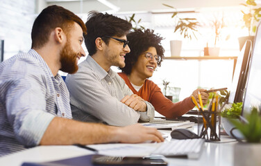 A diverse group of coworkers are gathered around a computer, engaging in a collaborative discussion...