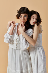 Two young women wearing white summer dresses embrace against a beige backdrop. Lesbian couple