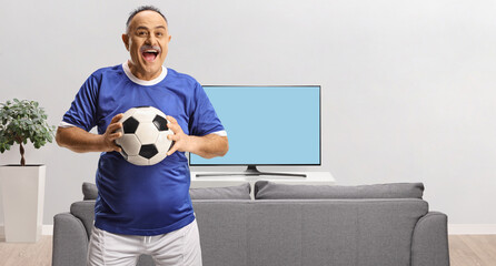 Cheerful mature man in a football jersey holding a ball at home