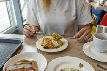 A woman dressed in a casual white shirt sits at a table and enjoys a piece of pastry, using a knife and fork to cut and savor the dish. The table is set with various plates and a cup of coffee.