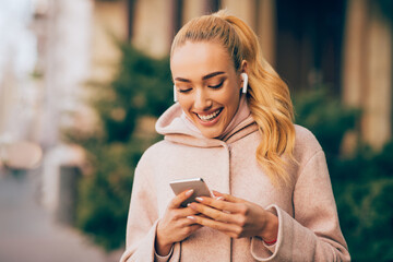 A young woman with blonde hair and a pink hoodie smiles while looking at her smartphone in an outdoor setting. She is wearing earbuds and appears to be enjoying her time on her phone. - Powered by Adobe