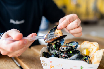 Casual dining experience: person eating mussels with a fork, served in a paper container with lemon and bread