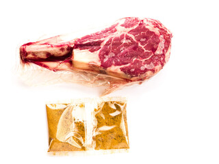 Fresh expensive tomahawk steak on the bone in a plastic vacuum package for freshness on white background and portion of herbs and spices. High quality beef meat product. Butcher craft. Food industry.