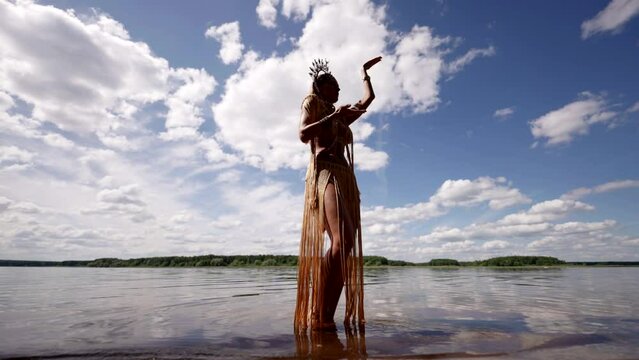 Against the backdrop of a picturesque lake and a blue sky with clouds, a woman in an exotic costume with a crown dances in the water, her movements are graceful and light.