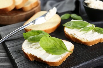 Pieces of bread with cream cheese and basil leaves on black table, closeup