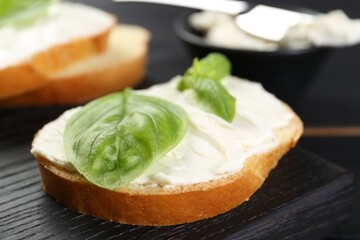 Piece of bread with cream cheese and basil on black table, closeup