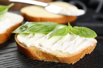 Piece of bread with cream cheese and basil on black wooden table, closeup