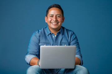 a Hispanic man in a blue casual shirt and jeans, looking happy while sitting with a laptop on a blue studio background