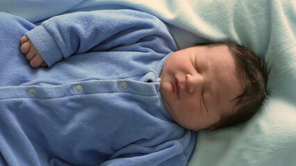 Close-up of a newborn baby sleeping in a blue onesie, nestled in a hospital bassinet, highlighting...