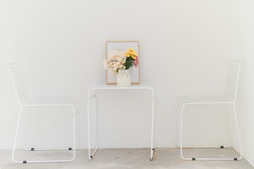 A minimalist white room featuring a small table with a floral arrangement, framed picture, and two white chairs, creating a clean and serene atmosphere.