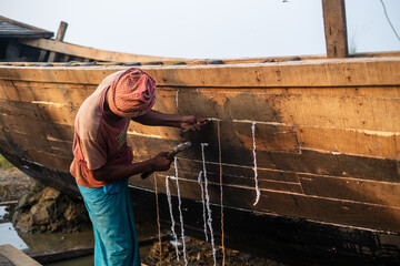 South asian young worker fixing gaps between woods of a newly constructed boat by ropes and hammer