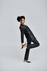A young African American woman poses in a black suit and heels against a grey background.