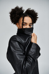 A stylish young African American woman poses in a black leather jacket against a grey background.