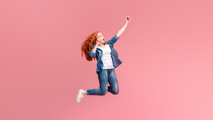 A spirited red-haired woman is caught mid-leap against a cheerful pink backdrop, her arm...