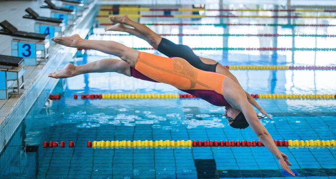 Two professional female swimmers taking track start position on the platform gracefully jumping and diving in a swimming pool. Sport competition concept.