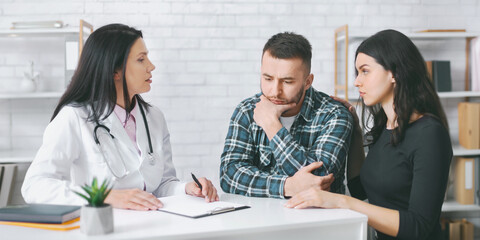 A doctor in a white coat is talking to a couple during a medical consultation in an office. The man is sitting with his head in his hands, while the woman is sitting next to him