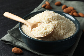 Fresh almond flour in bowl and spoon on dark textured table, closeup