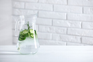 Refreshing cucumber water with rosemary in jug on white table against brick wall. Space for text