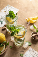 Two glasses of mojito cocktail or tonic with lemon, mint and ice on brown textured background. Refreshing summer drink