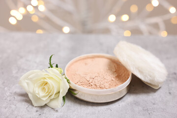 Face powder, puff applicator and rose flower on grey textured table against blurred lights, closeup