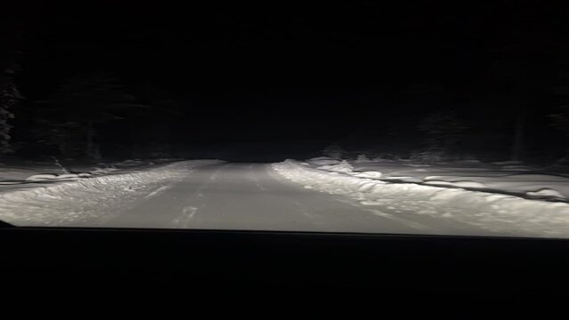 Vertical video, view of a snow covered road in the middle of the night in the forest around Rovaniemi, Lappland, Finland. White dry snow everywhere, car lights beams, darkness around. 
