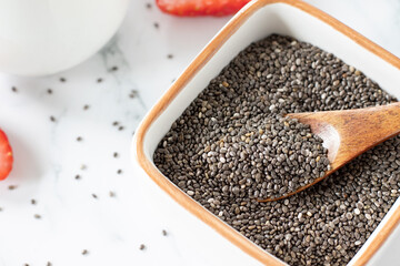 Raw organic chia seeds in a bowl with wooden spoon on white table with plant-based milk and strawberries for breakfast.