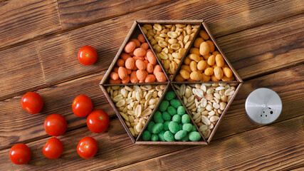 Six varieties of colorful roasted glazed peanuts in a craft box next to fresh tomatoes and a salt shaker on a wooden rustic background. Country bar party nuts. Photo. Copy space